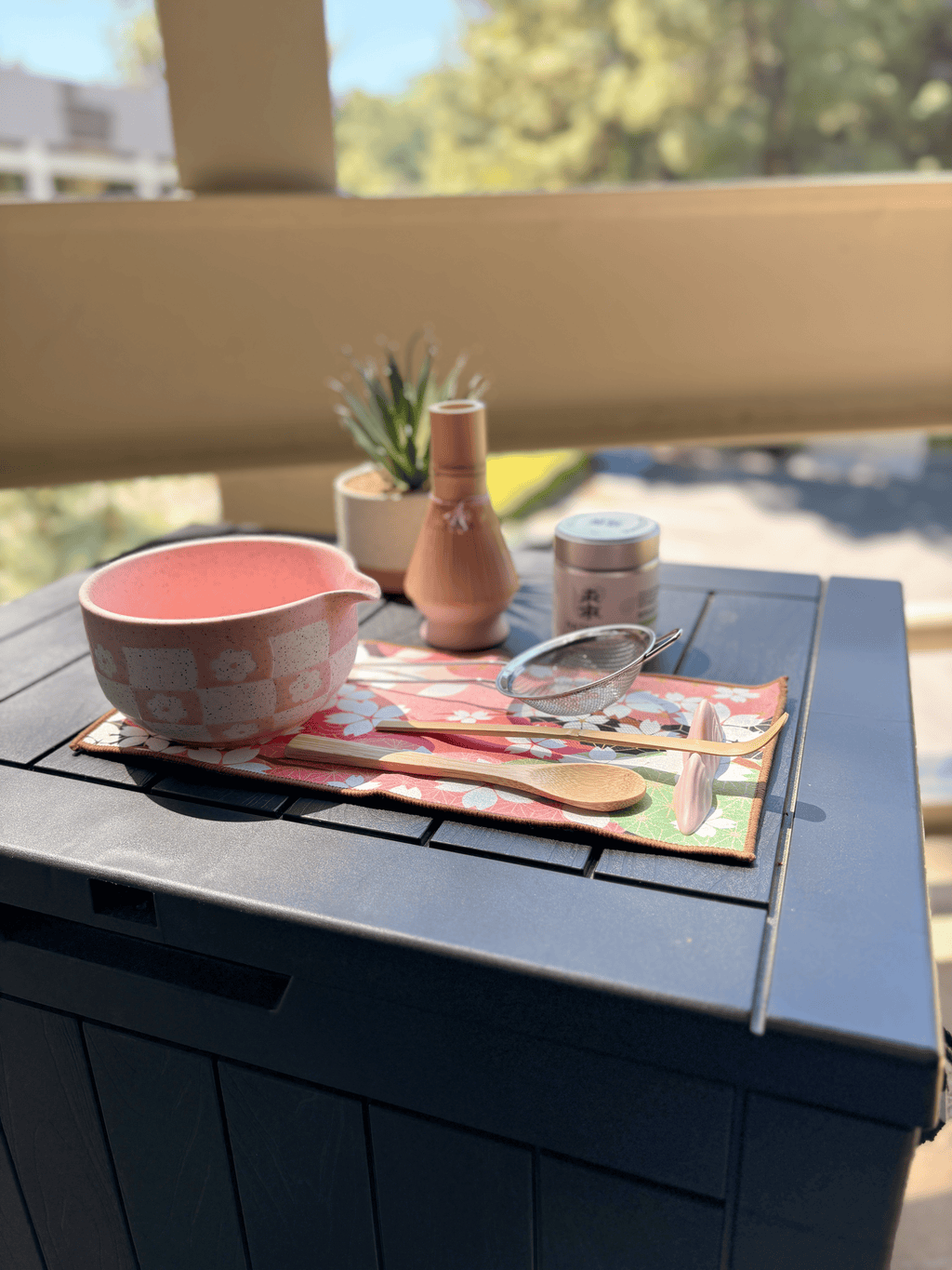 Full pink matcha tea set displayed on a black outdoor table, featuring a speckled chawan bowl, bamboo whisk with holder, scoop, wooden spoon, mesh sifter, matcha tin, and floral tea mat in natural sunlight.