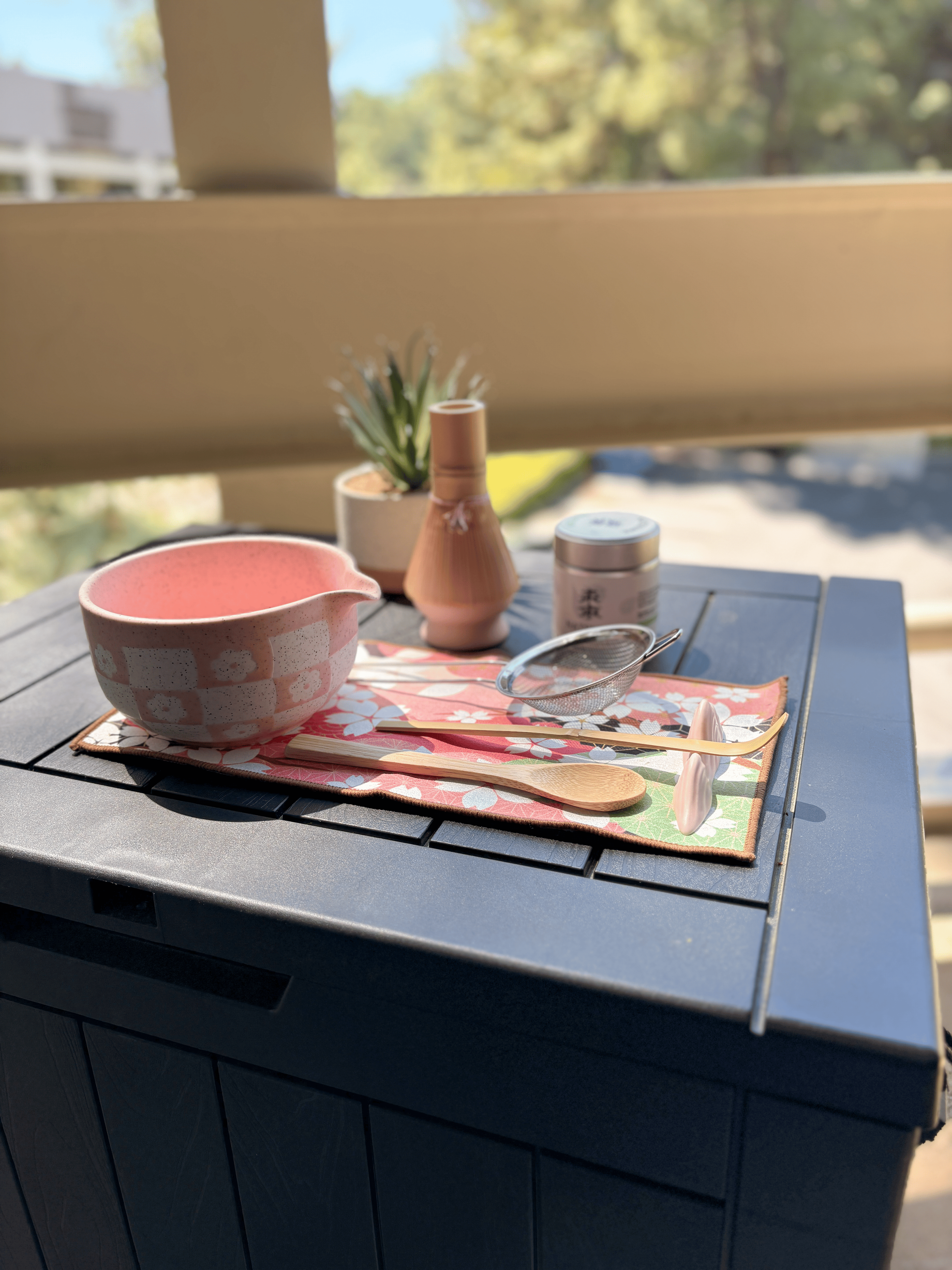 Full pink matcha tea set displayed on a black outdoor table, featuring a speckled chawan bowl, bamboo whisk with holder, scoop, wooden spoon, mesh sifter, matcha tin, and floral tea mat in natural sunlight.