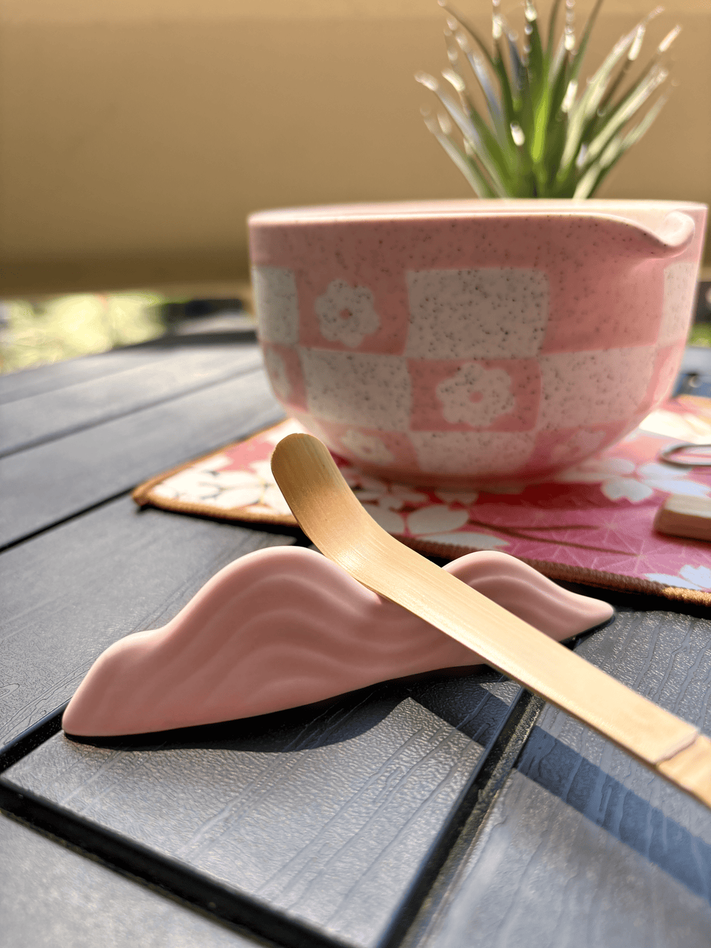 Partial view of a pink matcha tea set with bamboo scoop and floral mat on an outdoor table.