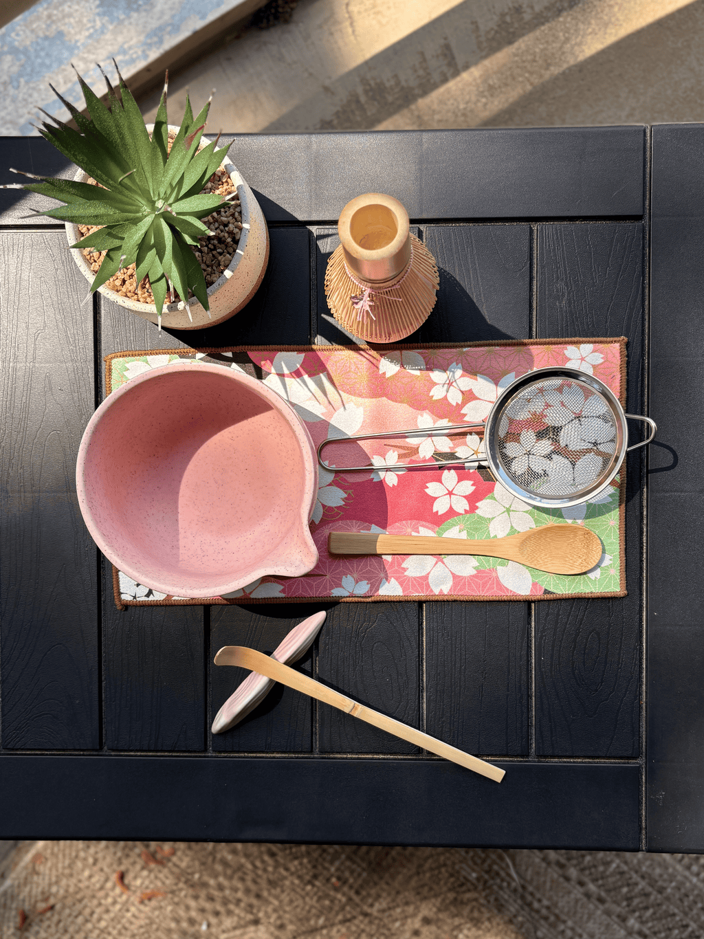 Top view of a pink matcha tea set featuring a speckled chawan bowl with pouring spout, bamboo whisk and scoop, wooden spoon, mesh sifter, and floral tea mat on a black table outdoors.