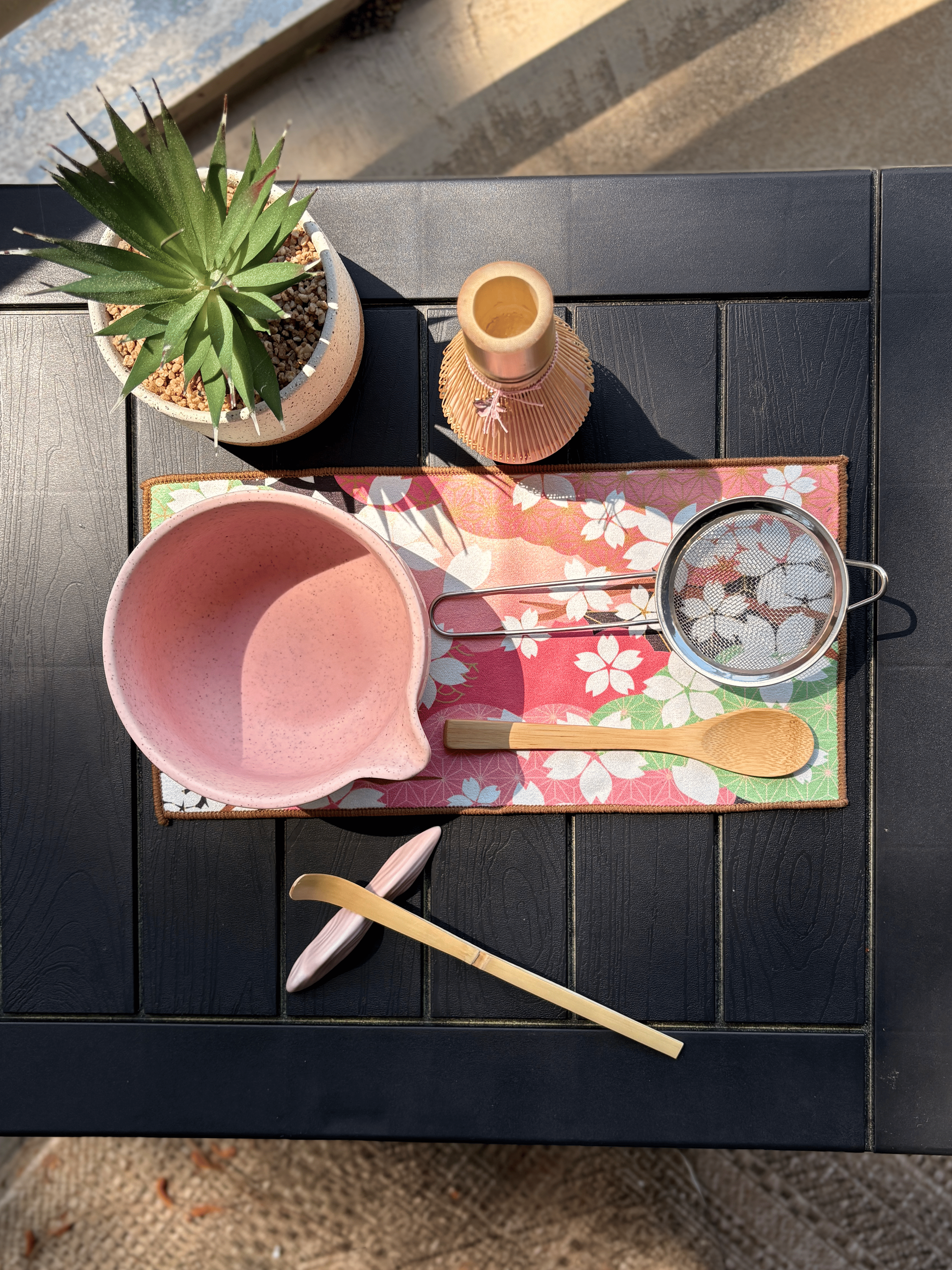 Top view of a pink matcha tea set featuring a speckled chawan bowl with pouring spout, bamboo whisk and scoop, wooden spoon, mesh sifter, and floral tea mat on a black table outdoors.