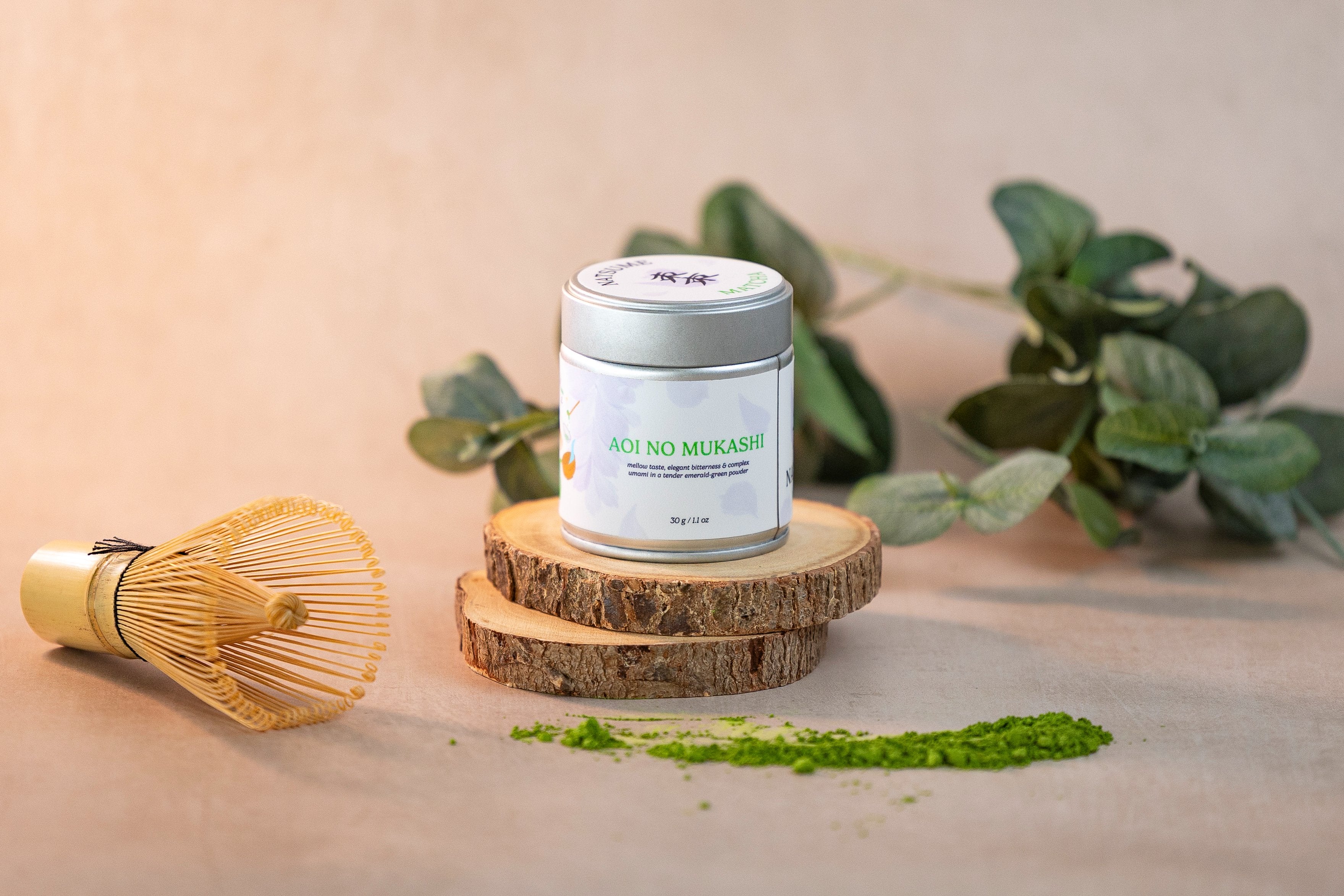 Natsume Matcha powder canister with a whisk on a wooden surface with green leaves in the background