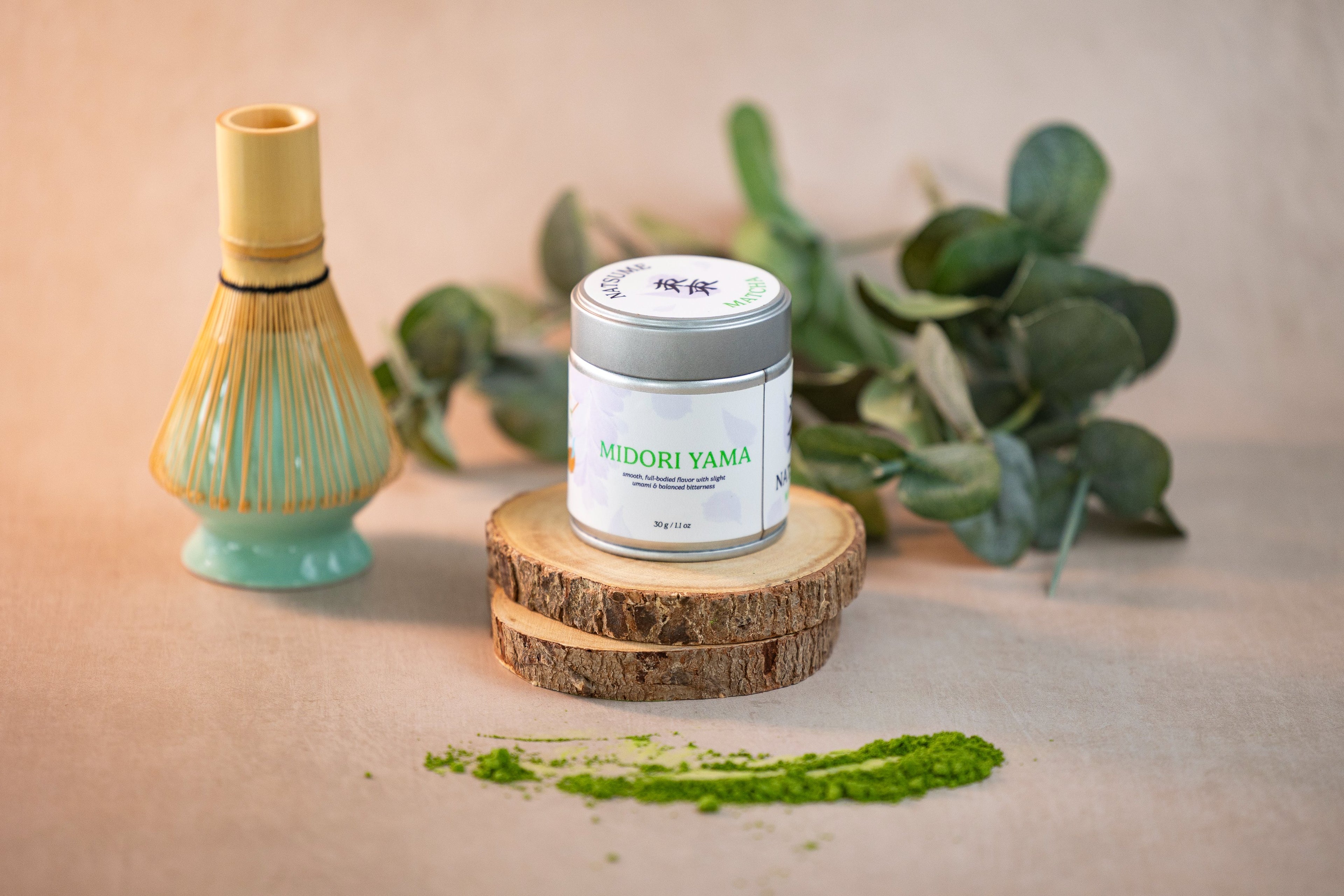 Matcha container and whisk on a wooden block with green leaves in the background