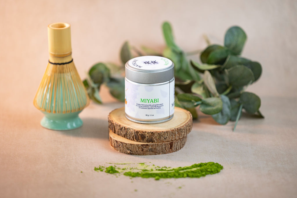 Natsume Matcha Miyabi product on a wooden stand with matcha powder and a whisk in the background