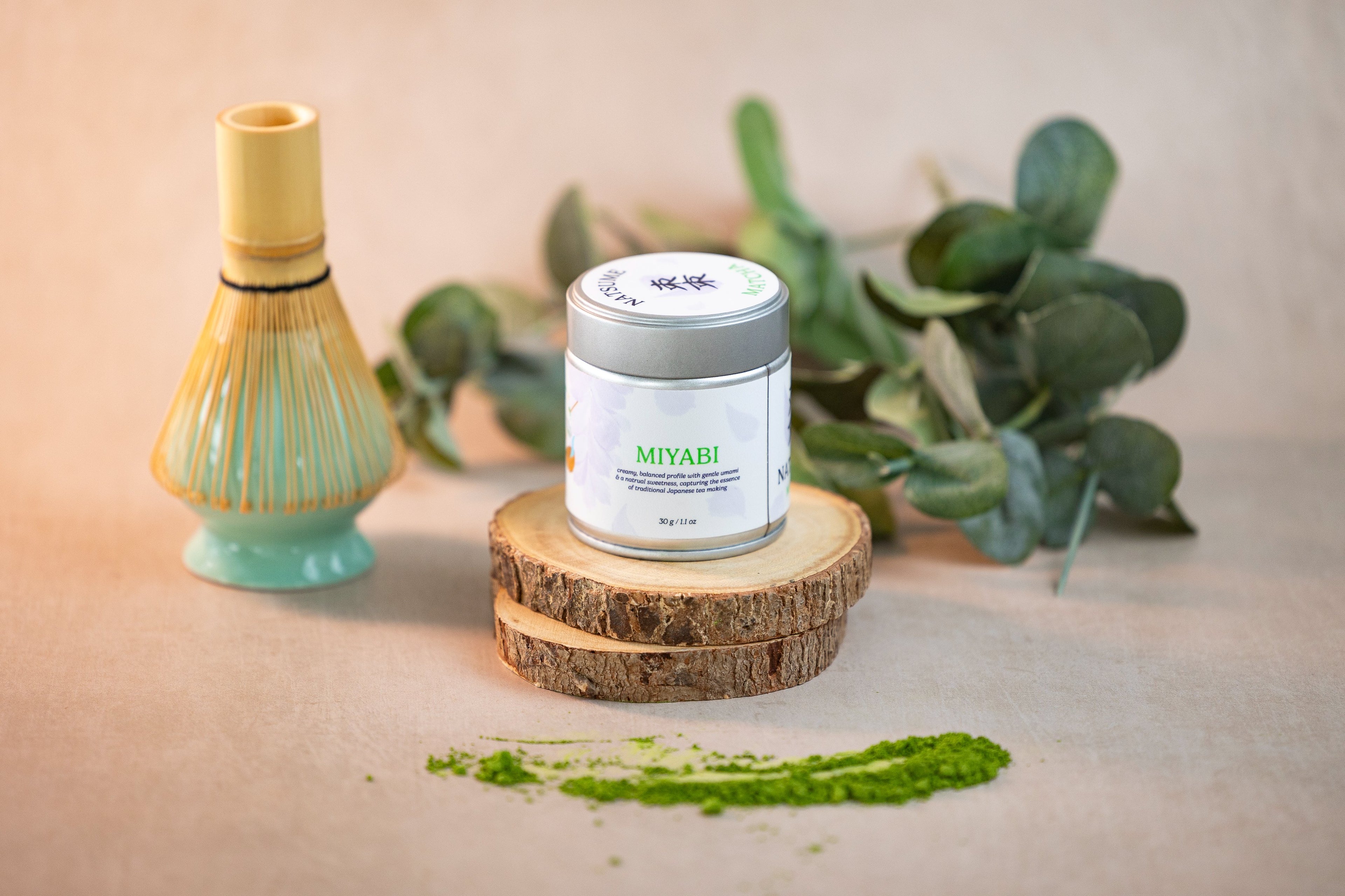 Natsume Matcha Miyabi product on a wooden stand with matcha powder and a whisk in the background