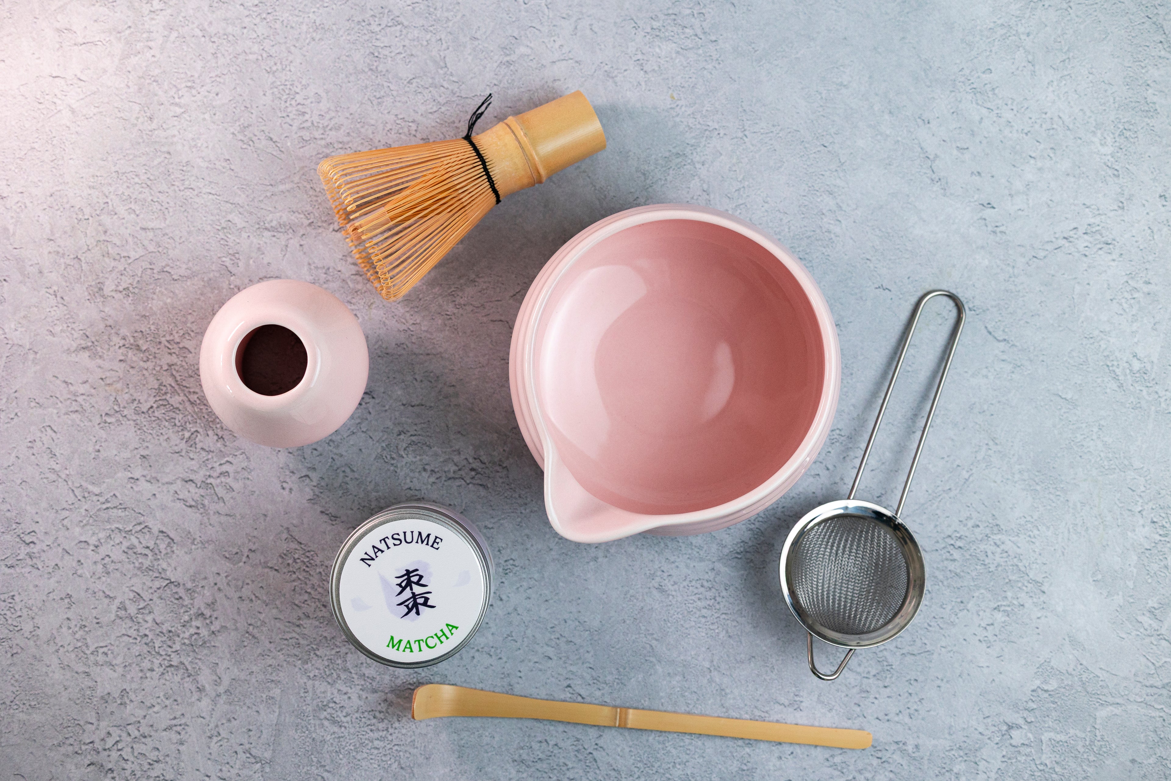 Matcha Tea-making accessories including a pink bowl, whisk, scoop, and container on a gray surface.