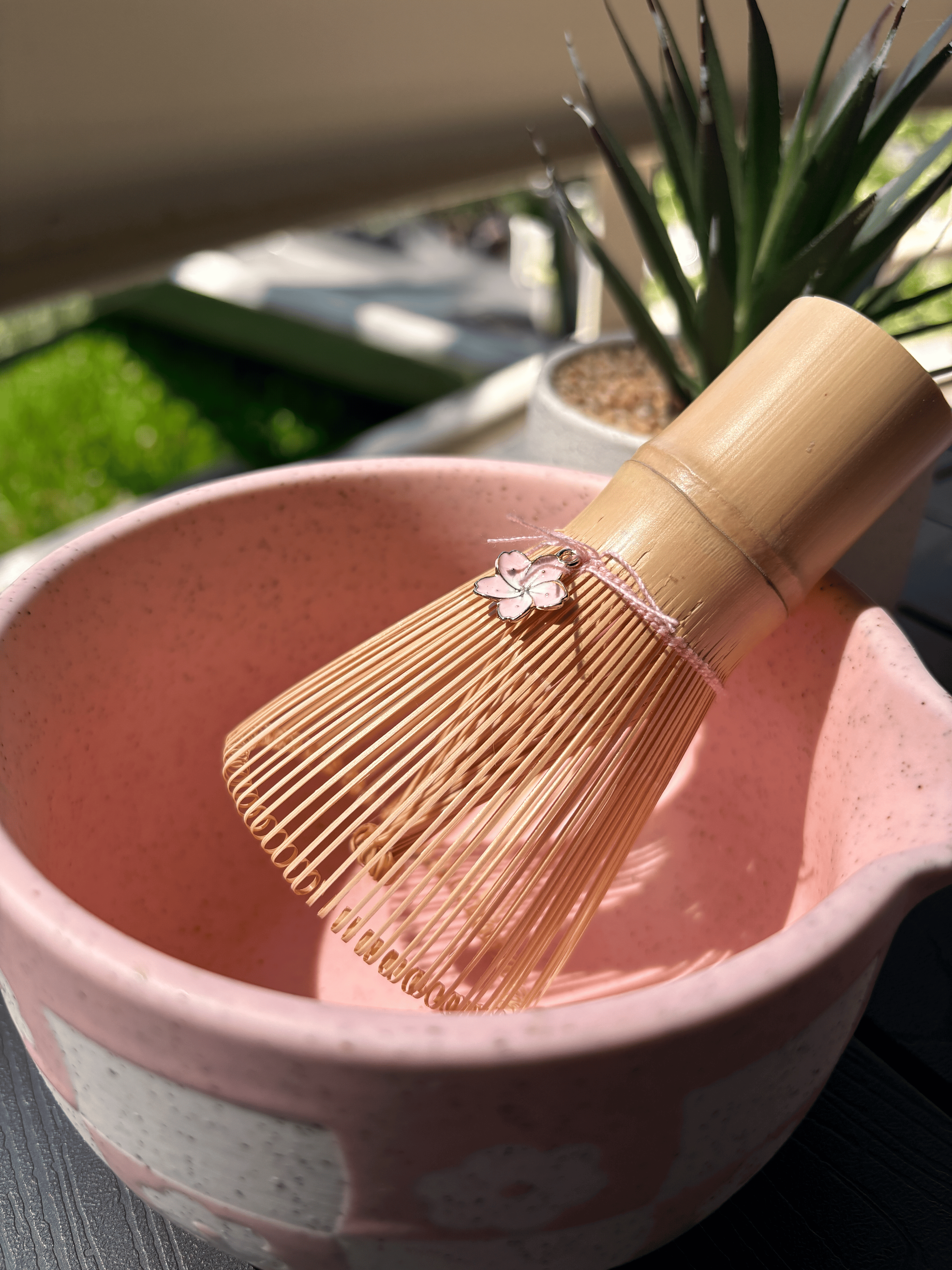 Close-up of a bamboo matcha whisk resting inside a pink speckled chawan bowl with a small cherry blossom charm, set outdoors in natural sunlight.