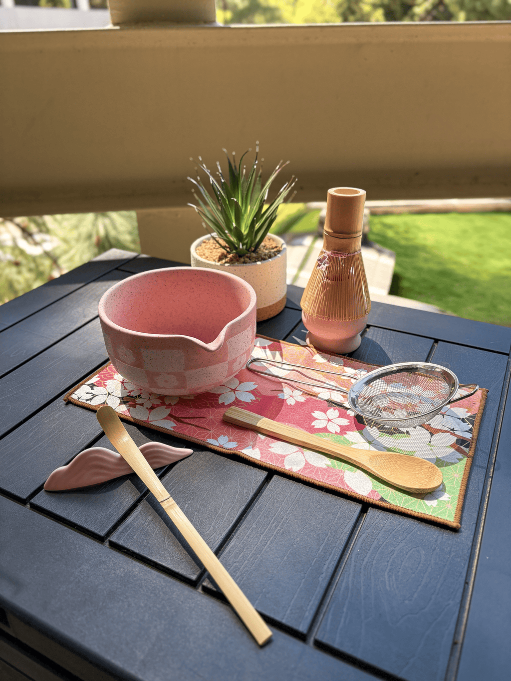 Top view of a pink matcha tea set featuring a speckled chawan bowl with pouring spout, bamboo whisk and scoop, wooden spoon, mesh sifter, and floral tea mat on a black table outdoors.