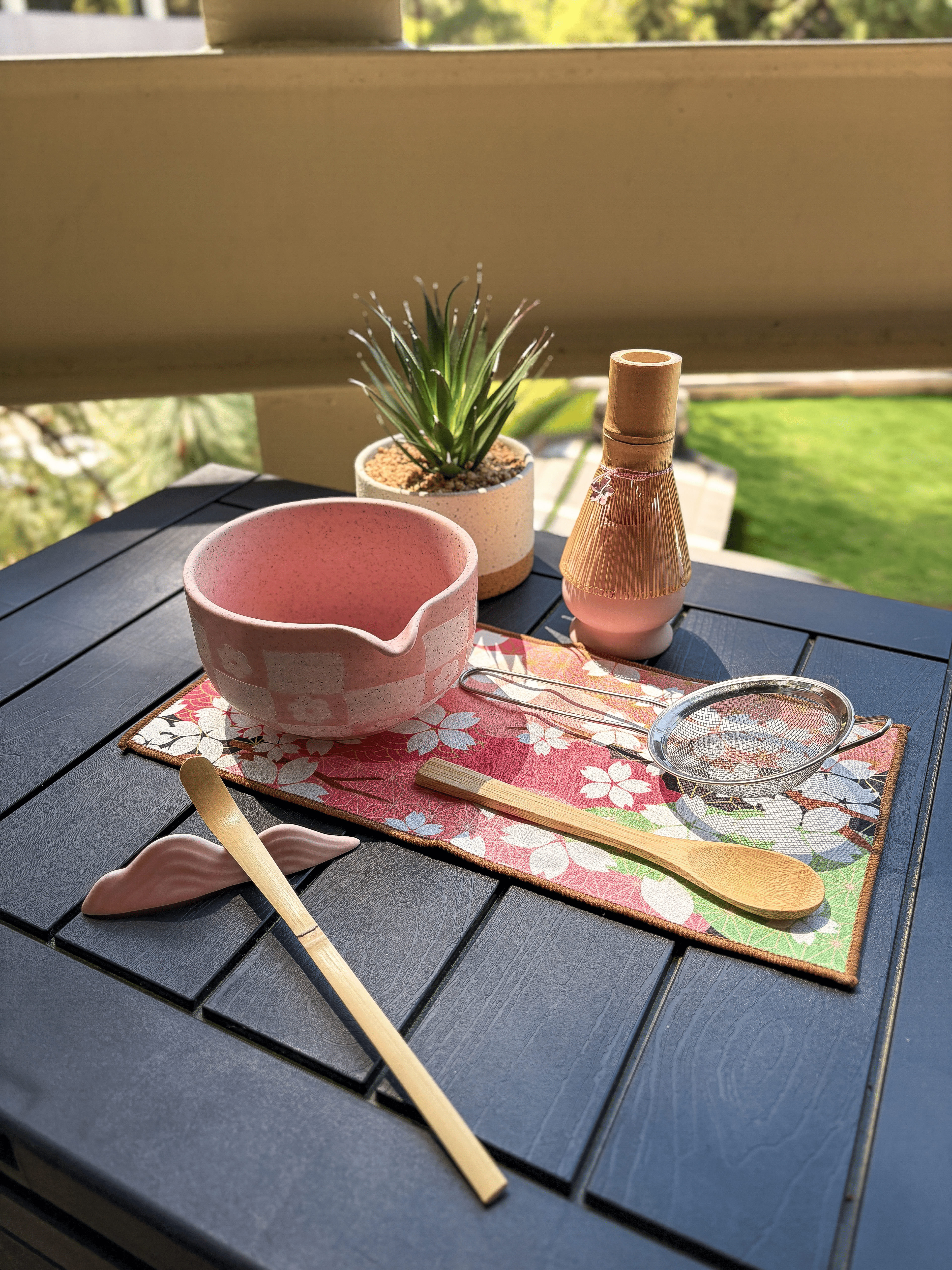 Top view of a pink matcha tea set featuring a speckled chawan bowl with pouring spout, bamboo whisk and scoop, wooden spoon, mesh sifter, and floral tea mat on a black table outdoors.